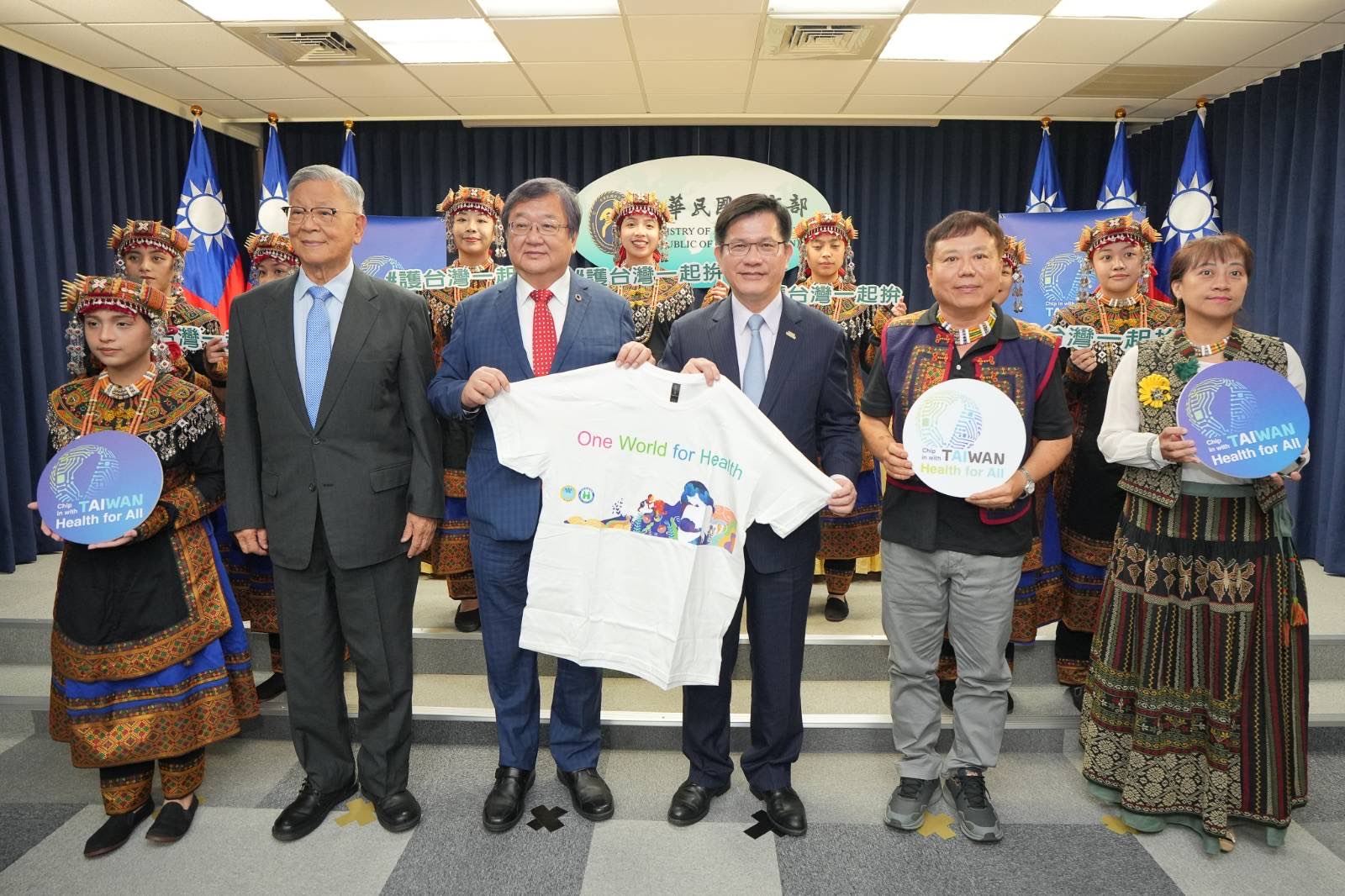 外交部長林佳龍(右三)及衛生福利部長邱泰源(左三)展示本年世衛行動團T-shirt,並與將隨團赴日內瓦的吳運東無任所大使及Puzangalan希望合唱團團員們合照。